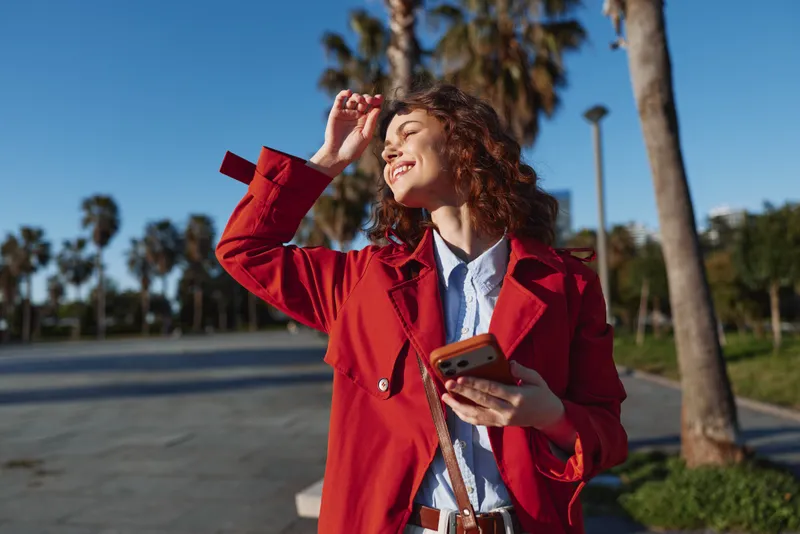 Smiling woman with red coat outdoors, sunlight on her face, palm trees in the background. Fashion lifestyle portrait emphasizing joy, warmth, and carefree confidence.