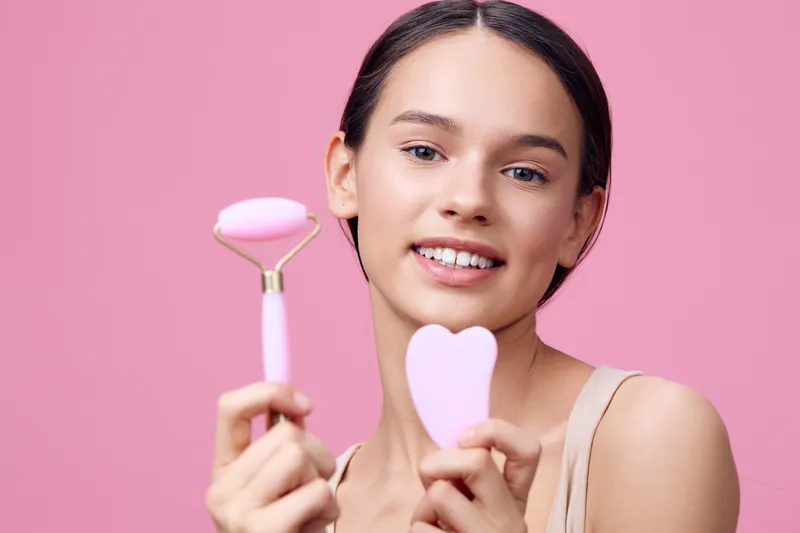 Young woman showcasing facial skincare tools on a pastel pink background, promoting healthy skin routines with a joyful expression