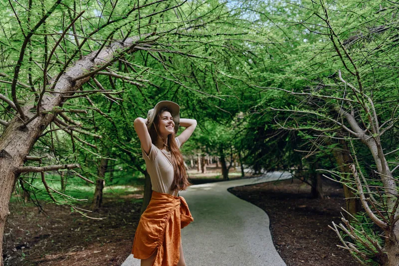 Woman on a winding path under dense trees in a park, smiling and relaxed while wearing a hat and casual dress, nature lifestyle portrait capturing joyful outdoor leisure, sunlight and greenery.