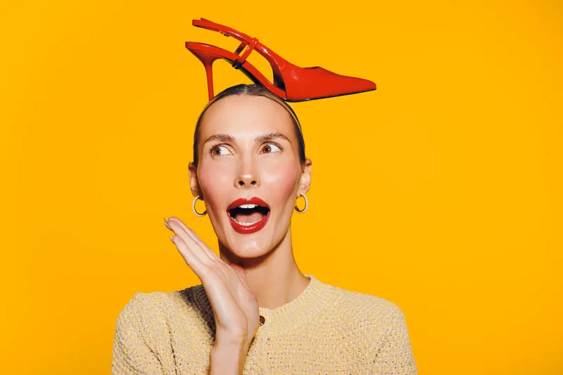 A surprised woman with red lipstick and hoop earrings holds a red high heel shoe on her head against a bright yellow background. Concept of fashion, fun, and creativity.