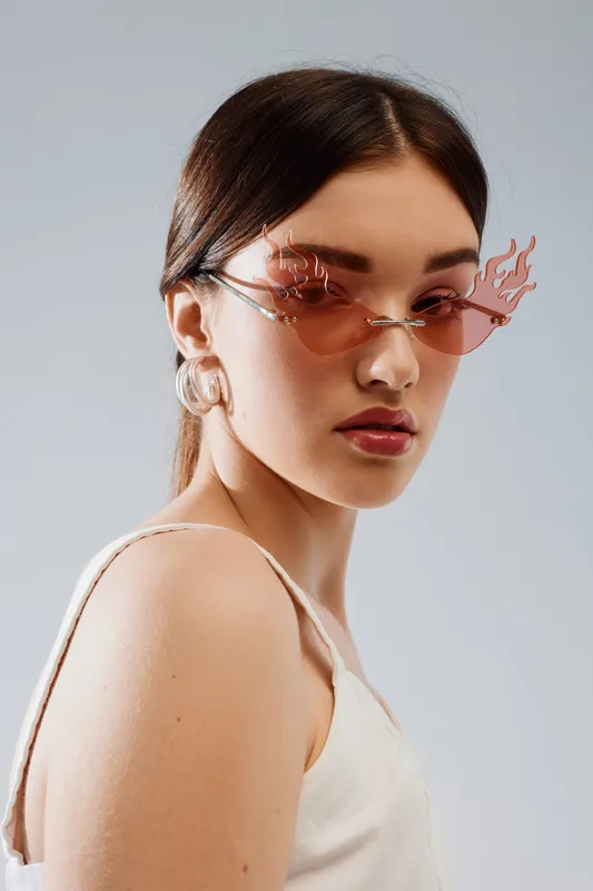 Young woman in trendy flame-shaped sunglasses and white top poses against a neutral background. Concept of beauty fashion and modern style.