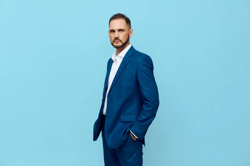A professional man in a blue suit stands against a solid blue background, exuding confidence and focus. The isolated color backdrop highlights corporate style and calm determination for business