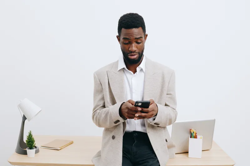 Business man with dark skin using smartphone at office desk, modern workspace with laptop, plant, stationery, minimalistic white background, professional lifestyle concept.