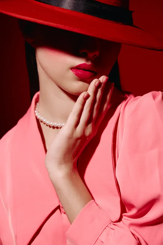Fashion woman in coral outfit with red hat partially covering her face poses with pearly beads and glossy lips. Creative beauty portrait themed on elegance and bold style.