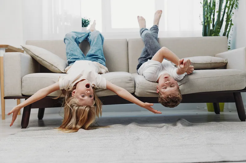 Two playful children lie upside down on a beige sofa, laughing and stretching arms in a bright living room. Concept of childhood joy, carefree moments, and sibling fun.