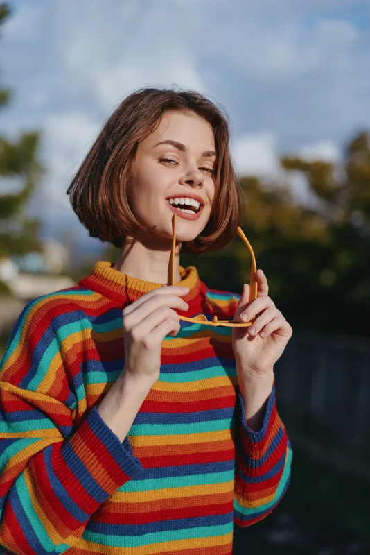 Woman smile sunglasses sweater portrait outdoors happy young female in colorful striped sweater holding sunglasses, short bob hair, cheerful casual style with natural sunlight in urban backyard.