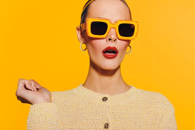 Stylish woman in yellow sunglasses and beige sweater poses with surprised expression on bright yellow background. Concept of fashion, style, and vibrant portrait.