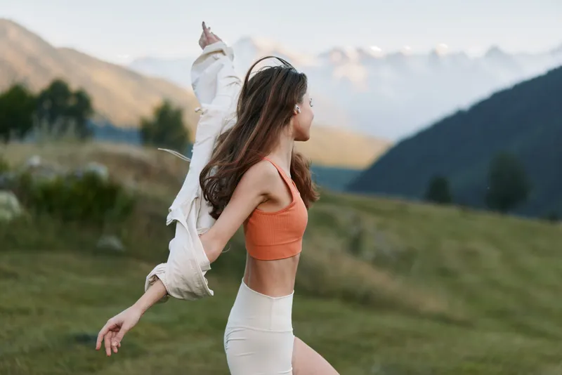 A joyful woman wearing an orange crop top and white pants enjoys a carefree moment outdoors, surrounded by rolling hills and distant mountains, radiating energy and freedom.