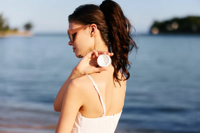 Portrait of a young woman by the seaside wearing a white dress and sunglasses, turning slightly away while holding a seashell against her shoulder.