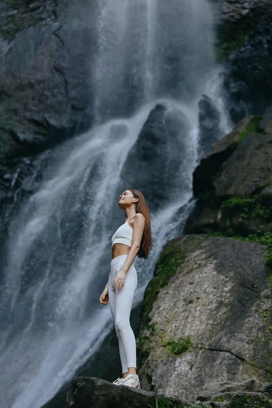 Woman standing near waterfall in white activewear, surrounded by rocks and greenery, showcasing a peaceful connection with nature and outdoor fitness.