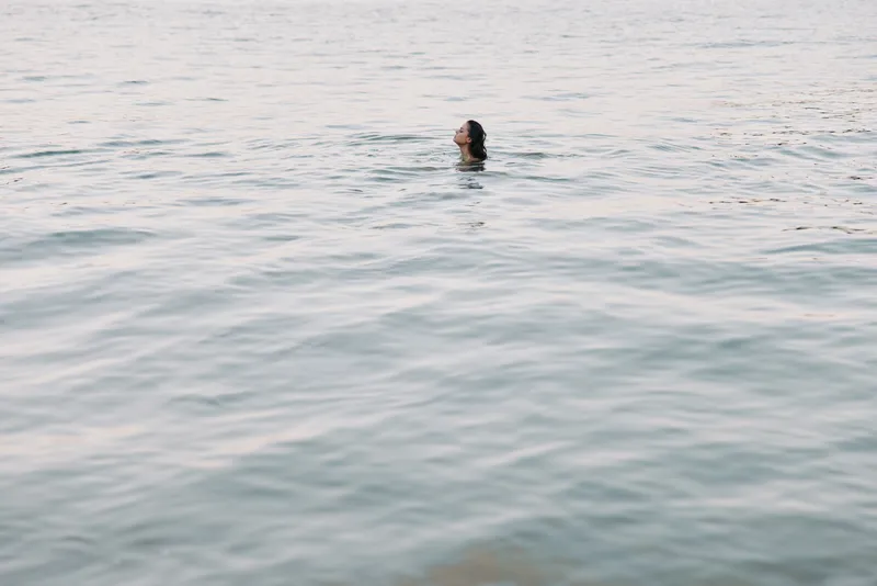 A lone swimmer rises from calm ocean water, head and shoulders breaking the glassy surface as soft light plays on gentle ripples and serene blue tones.