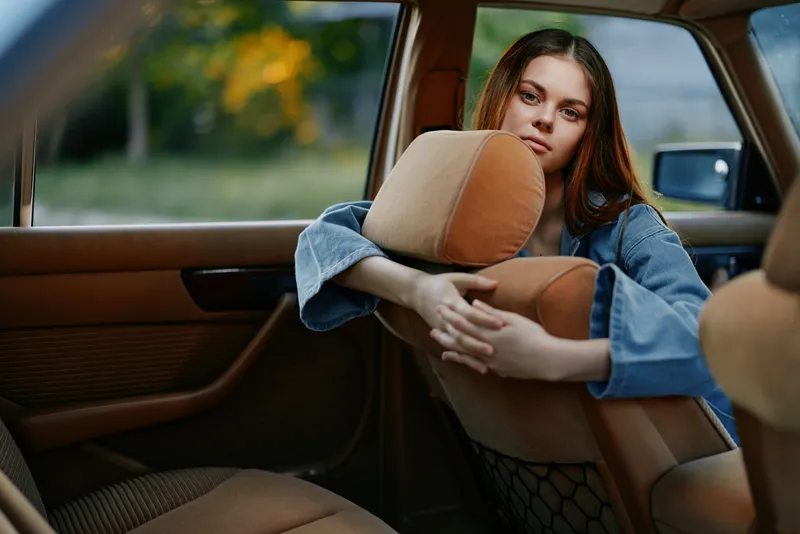 Young woman in a denim jacket sitting in a vintage car with a contemplative expression, surrounded by nature and warm evening light, evoking a sense of nostalgia.