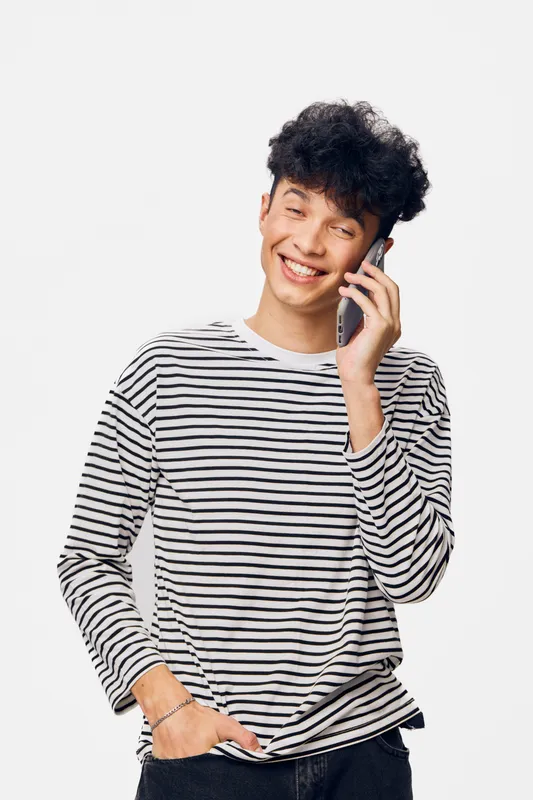 Smiling young man talking on smartphone wearing striped long sleeve shirt, casual style, curly hair, isolated on white background, cheerful and relaxed mood.