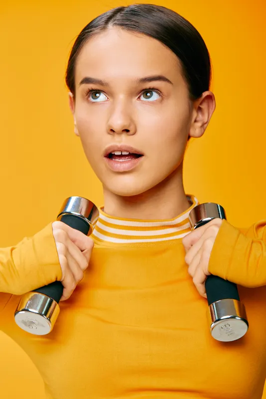 Athletic woman fitness enthusiast lifts dumbbells in a bright orange studio, showcasing strength, focus and healthy lifestyle during a dynamic workout.