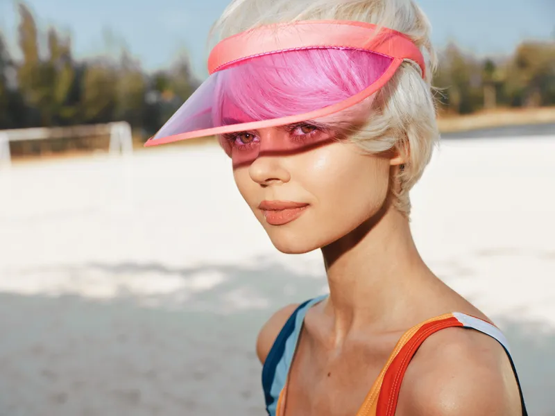 A stylish woman on a sunlit beach wears a pink visor and vibrant swimsuit, posing with soft light that highlights clear skin and a vibrant coastal vibe.