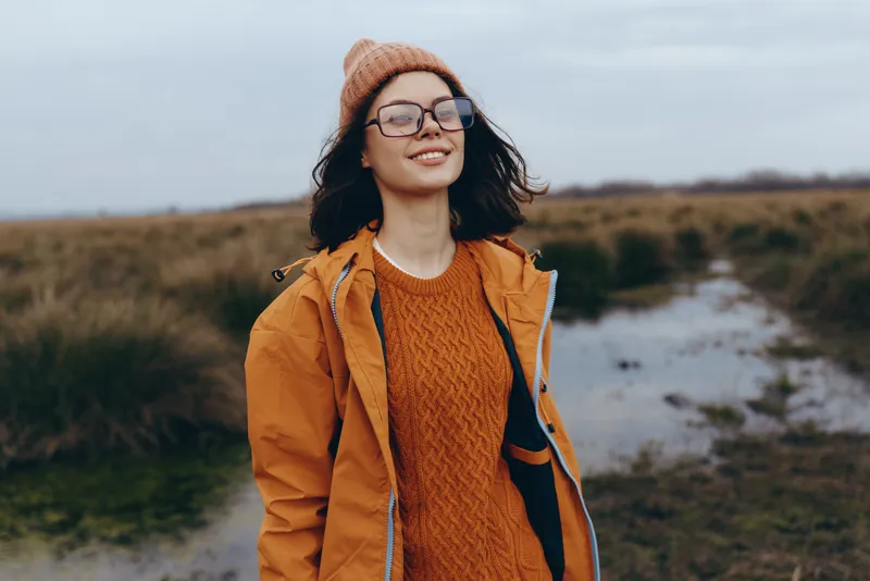 Smiling woman outdoors in orange jacket and knitted hat, wearing glasses, standing in a rural field. Lifestyle and autumn travel mood with soft evening light, calm and fresh air.