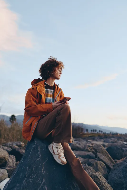 Woman lifestyle outdoors portrait, sitting on a rock with curly hair, wearing an orange jacket, relaxed expression, natural mountains and blue sky behind, calm travel mood