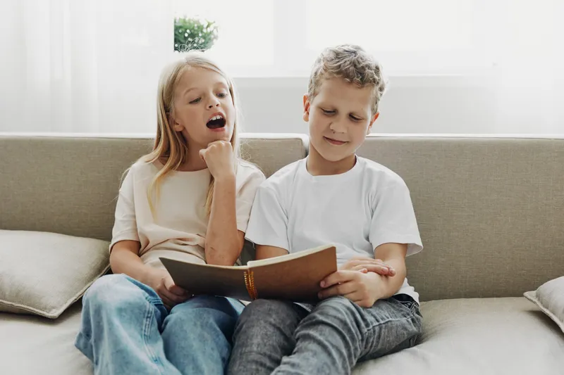 Two children sitting on a sofa reading a book together, girl yawning and boy focused, casual clothing in a bright living room. Concept of childhood, learning, and family moments.