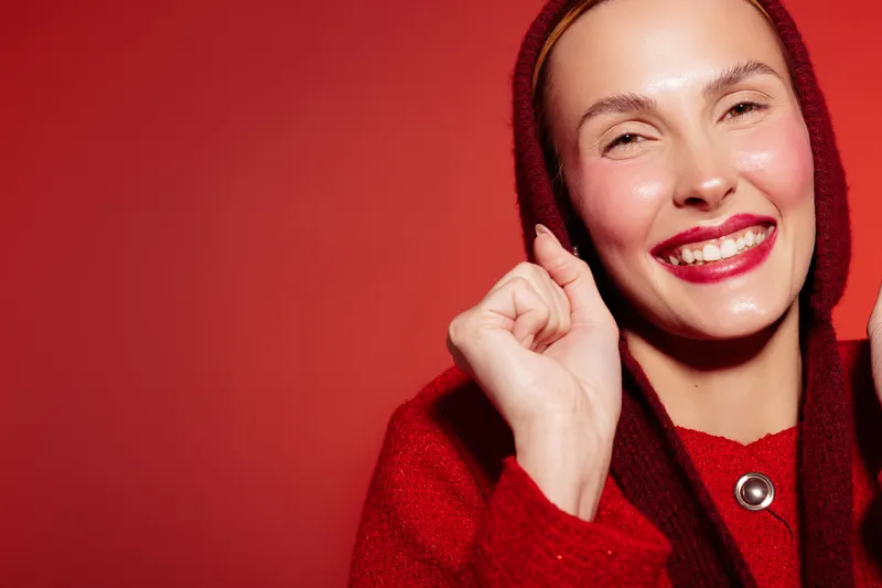 Smiling woman in red hoodie with bright lipstick poses against red background. Close-up portrait with natural skin glow and happy expression. Concept of fashion, beauty, and confidence.