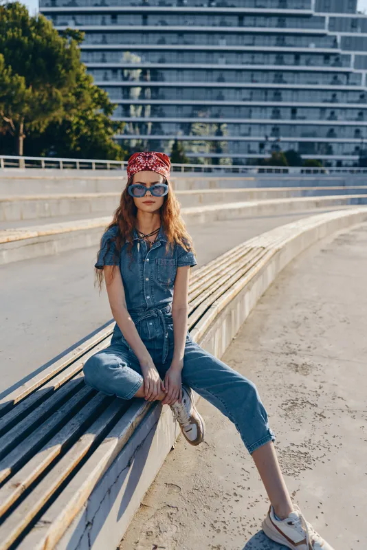 Lifestyle woman in modern boho-western denim street style with sunglasses and red bandana, captured in vibrant film color under urban sunlight.