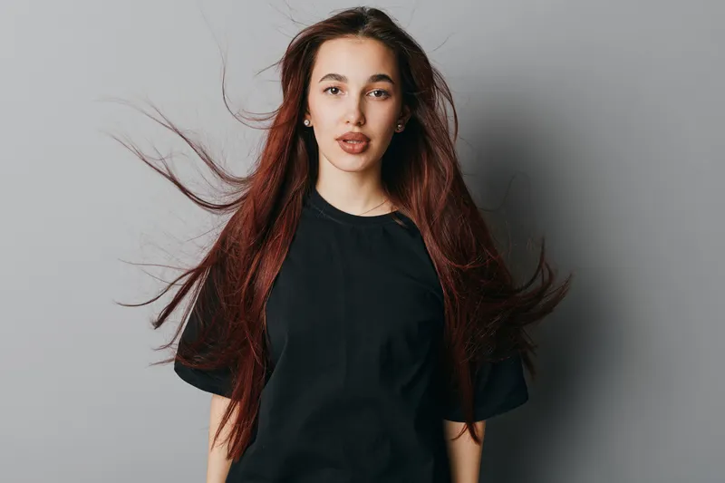 Young woman with long red hair and black t shirt standing against gray background with hair flowing and neutral expression looking at camera in studio portrait.