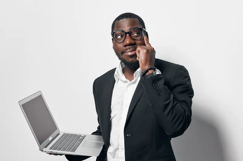 African American man with dark skin wearing glasses and black suit holding laptop and thinking with finger on temple on white background.