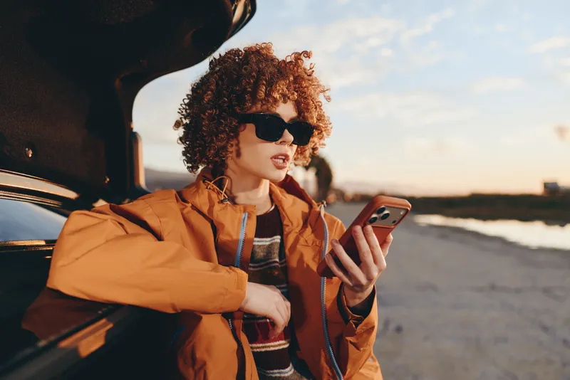 Woman with curly hair wearing sunglasses, orange jacket, holding smartphone, sitting in car by open door at sunset, road trip lifestyle, warm sunlight, outdoor travel portrait