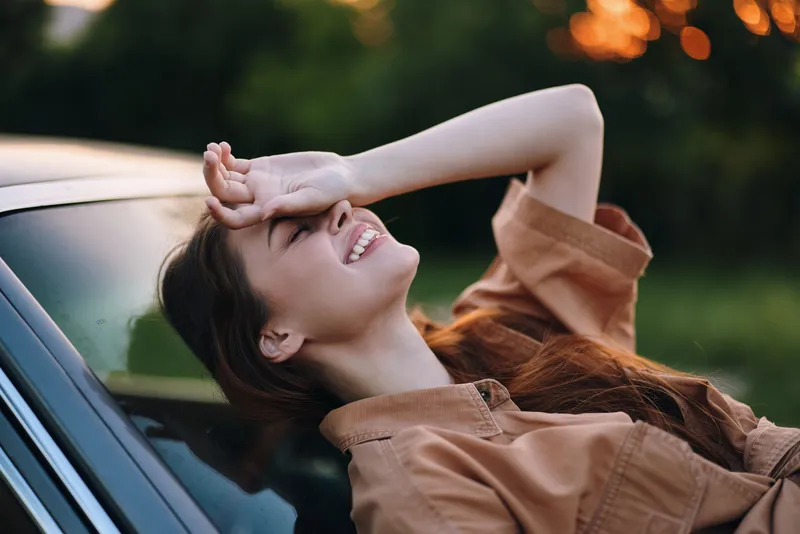 Cheerful young woman enjoying sunset outdoors, relaxing on car hood, wearing casual brown shirt, conveying happiness and peace in nature's warm hues.