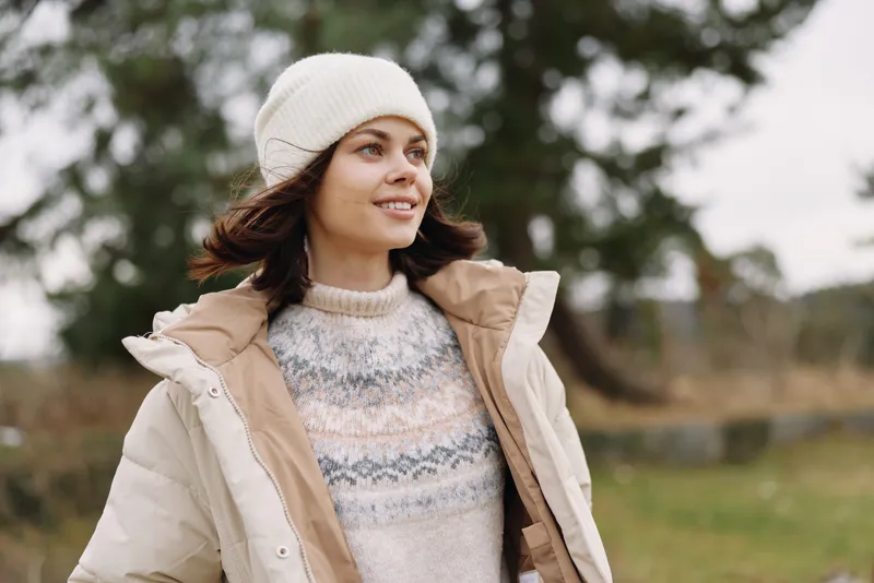 Woman in a warm beige jacket and knit sweater stands outdoors with a beanie, enjoying a breezy countryside scene in cool weather. A relaxed pose conveys seasonal fashion and comfort.