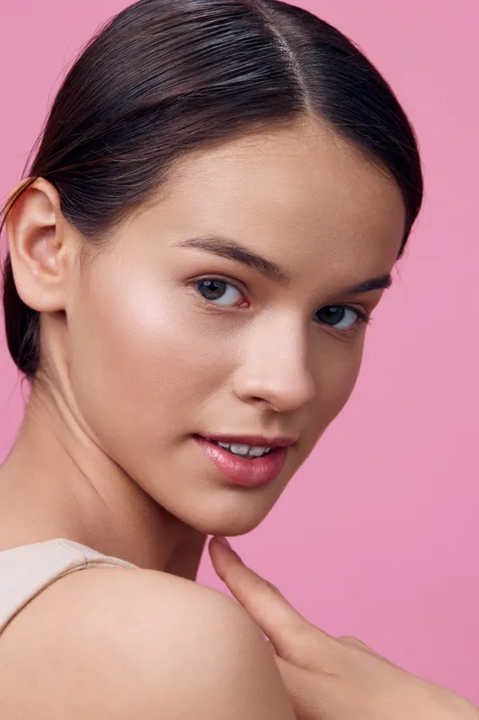 Portrait of a young woman with clear skin and natural makeup against a pink background, exuding confidence and elegance