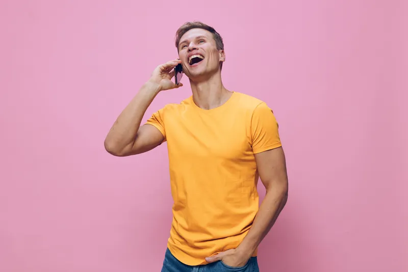 Smiling man talking on the phone cheerful casual young male wearing yellow t-shirt, blue jeans standing against pink background, demonstrating happiness and relaxation in a lifestyle portrait. People 