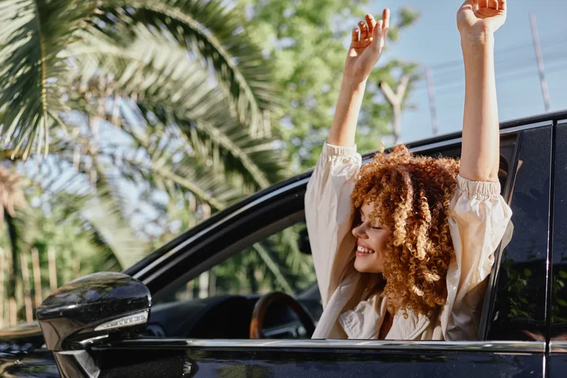 Smiling woman with curly hair enjoys sunny day in a car with tropical palm trees in the background, expressing happiness and freedom outdoors.
