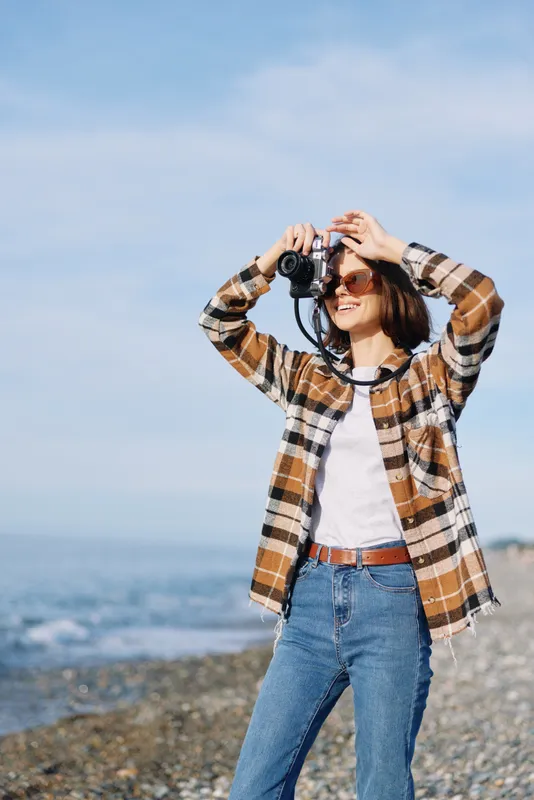 Casual woman with a camera on a breezy beach, scanning the coastline and capturing candid moments in daylight, wearing a plaid shirt and jeans by the sea