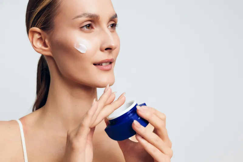 Young woman with clear skin applies moisturizing cream on her cheek while holding a blue jar in a bright studio, showing skincare routine and self-care concept.