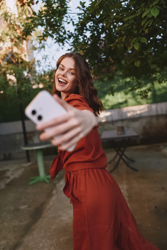 Smiling woman in a red dress takes a selfie in an outdoor park, sunlight streaming through trees, creating a vibrant lifestyle mood, casual, energetic moment, candid and warm