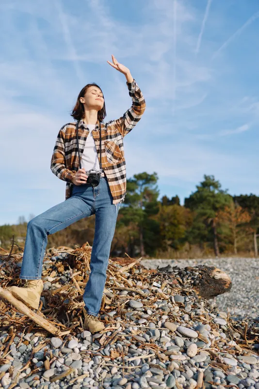Outdoor woman posing with a camera on a rocky shoreline under a clear blue sky, casual flannel and jeans, capturing nature and landscape with confident posture.