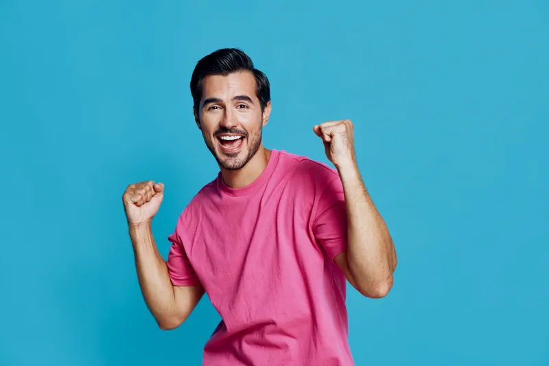 Cheerful young man celebrating success with raised fists, wearing a pink t-shirt, smiling joyfully against a bright blue background. People lifestyle concept.
