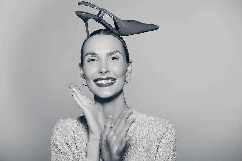Smiling white woman with pale skin wearing sweater and a high heel shoe on her head poses against grey background. Concept of humor, fashion, and creativity.