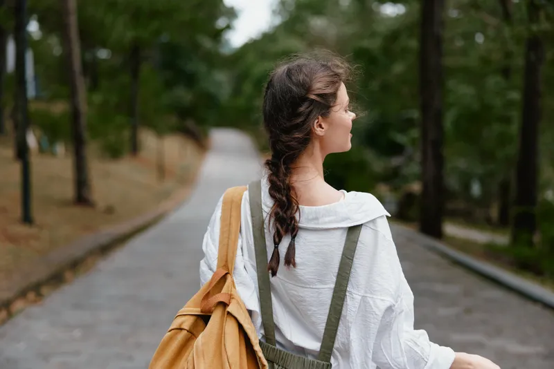 Young woman walking in a park, wearing a casual white shirt and green overalls, showcasing a carefree emotion with a scenic natural background