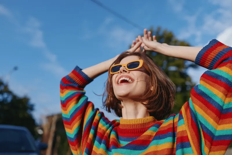 Woman smile sunglasses rainbow sweater joyful outdoor portrait happiness captured in sunlight, young woman in colorful casual fashion expressing carefree energy and radiant cheerful expression.