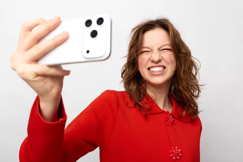 Young woman in red sweater takes a selfie with a white smartphone, smiling with eyes closed. Studio portrait on white background, concept of social media and self-expression.
