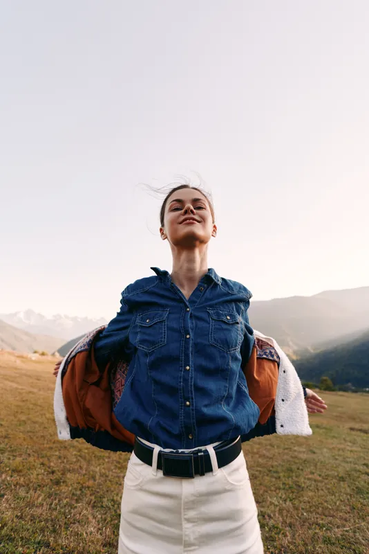 Woman with smile and denim shirt outdoors in nature on a grassy field with mountains in background, arms open showing freedom and joy, casual jacket and relaxed portrait.