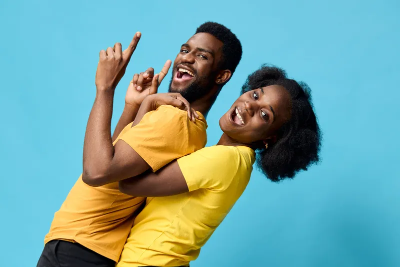 Happy diverse couple in matching yellow t shirts, joyfully celebrating against a bright blue background, showcasing fun and connection in a vibrant atmosphere