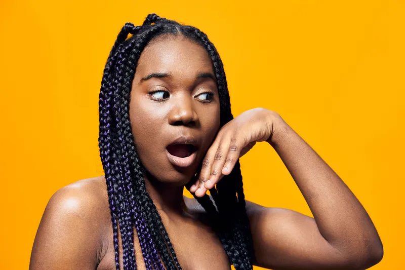 Surprised young woman with braids against a vibrant yellow background, expressing excitement and curiosity with her hand on her face