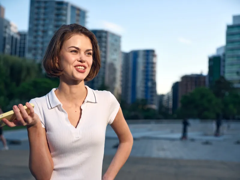 A cheerful woman stands outdoors in an urban park setting, smiling and speaking with a confident posture, conveying a friendly and approachable atmosphere.