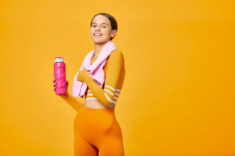 Woman in bright orange athletic wear standing with a towel over her shoulders and a pink water bottle in hand, ready for a workout, exuding energy, health and a bold fitness lifestyle against a