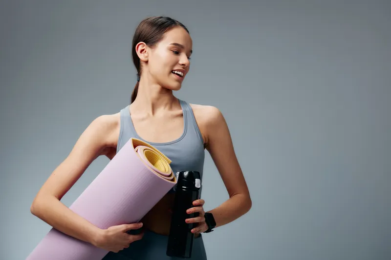 Woman in athletic wear holds a rolled yoga mat and a water bottle, ready for workout in a bright studio. Her hair is tied back, smartwatch on her wrist, and a calm smile complements the energetic