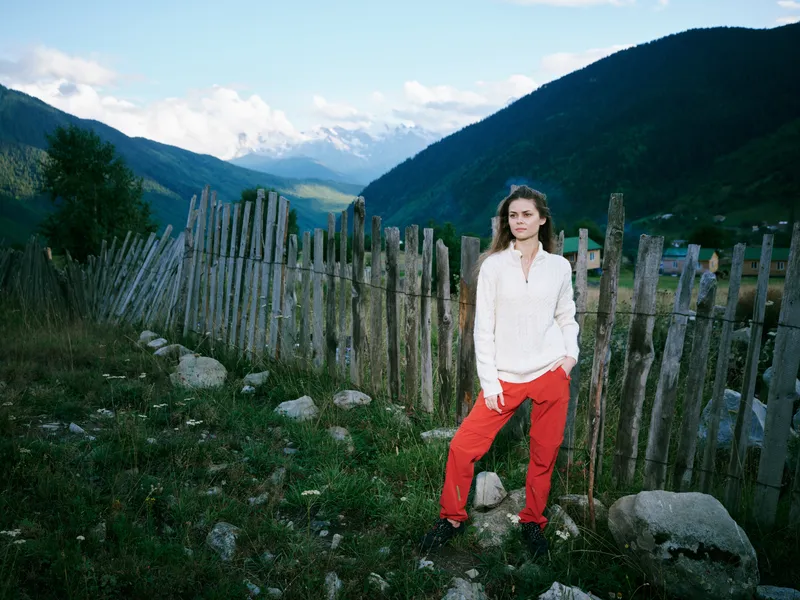 Woman in a white shirt and red pants stands beside a rustic wooden fence in a mountain landscape, outdoor scene showcasing nature, travel vibe, casual style, and rugged scenery