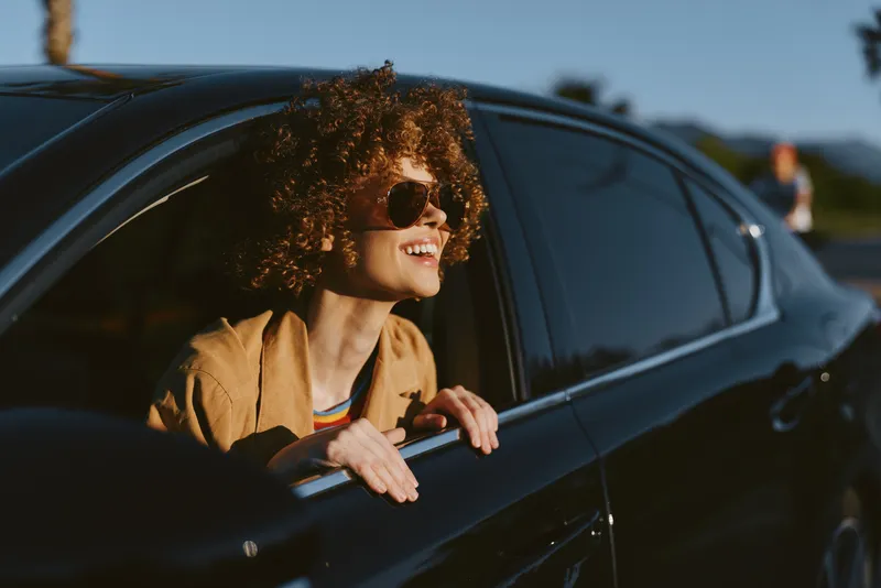 A young woman with curly hair leans out of a sleek car window, wearing sunglasses and a light jacket, enjoying a sunny drive. The retro color palette adds a film vibe to this candid moment.