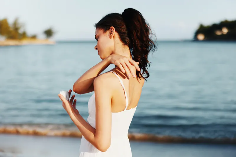 Sunlit beach scene featuring a young woman in a white dress, standing by tranquil water, gazing toward the horizon, while she gently touches her shoulder and brushes her hair aside.